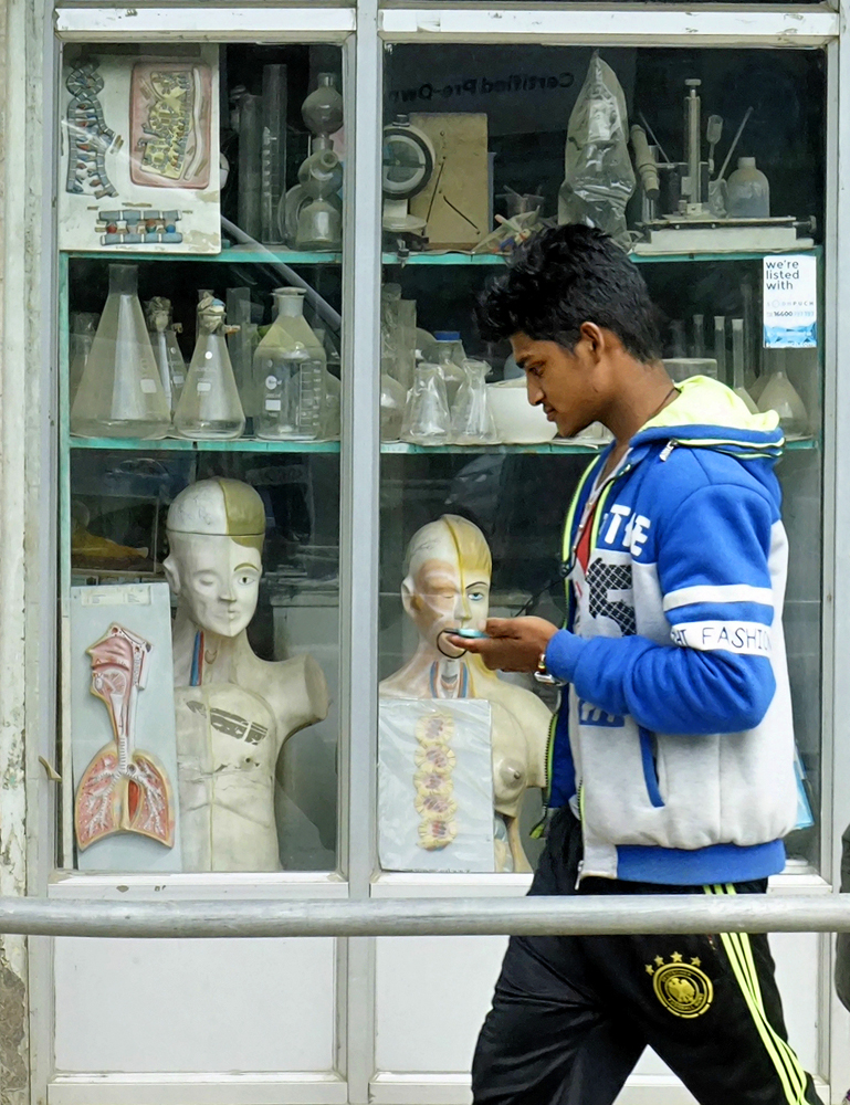 Street photography, storefront, shop window, human anatomy, anatomical model, laboratory glassware, scientific equipment, man with cell phone, passer-by, Nepal